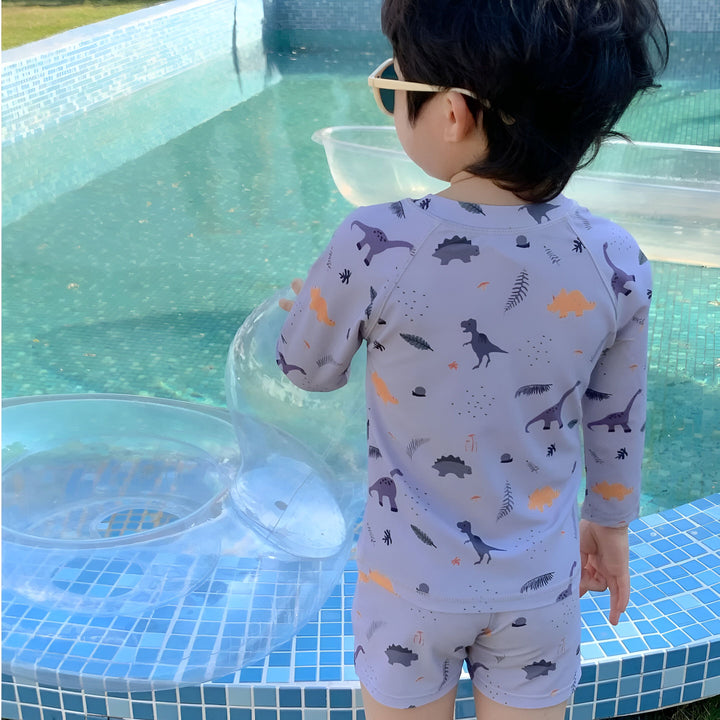 Un garçon en maillot de bain beige à motif de palmiers, avec des lunettes de soleil, près d'une piscine, levant la main. Bouée de sauvetage à rayures bleues et blanches visible. Style de vie estival, enfant à la piscine.
