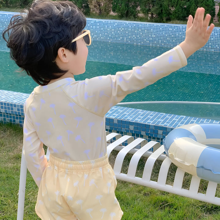 Un garçon vu de dos près d'une piscine, portant un maillot de bain à motifs de dinosaures et des lunettes de soleil. Il tient une bouée transparente. Ambiance estivale et ludique.