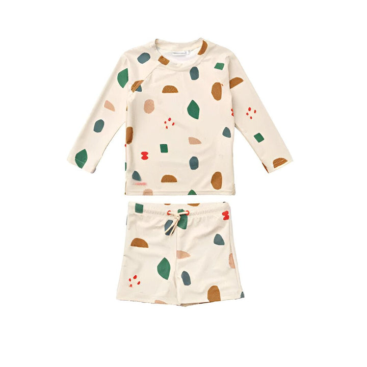 Garçon en maillot de bain imprimé avec un ballon de plage vert et blanc, posant devant un mur de pierre. Ambiance estivale et joyeuse. Mode enfant plage.