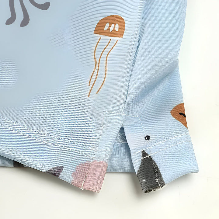 Gros plan sur un vêtement pour enfant bleu clair avec des motifs marins : méduses, hippocampe, baleine et bulles. Fermeture éclair visible.