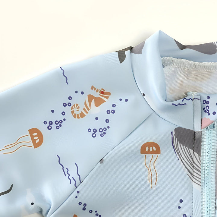 Détail d'un vêtement bleu clair imprimé avec des motifs marins (méduses, pieuvres). L'ourlet est visible avec des coutures blanches apparentes.