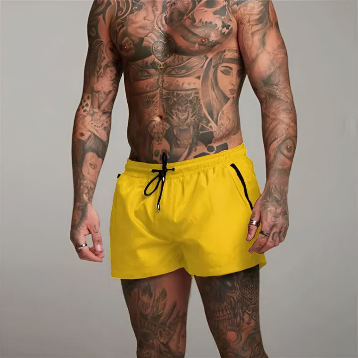 Homme musclé de dos portant un short de bain bordeaux avec des tatouages sur le bras et le dos. Vêtements de plage pour homme, style décontracté.