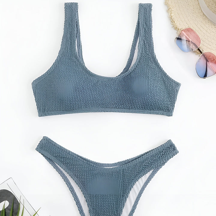 Image en gros plan d'une femme portant un maillot de bain rose texturé contre un ciel bleu clair. Collier doré délicat visible. Style de mode estival.