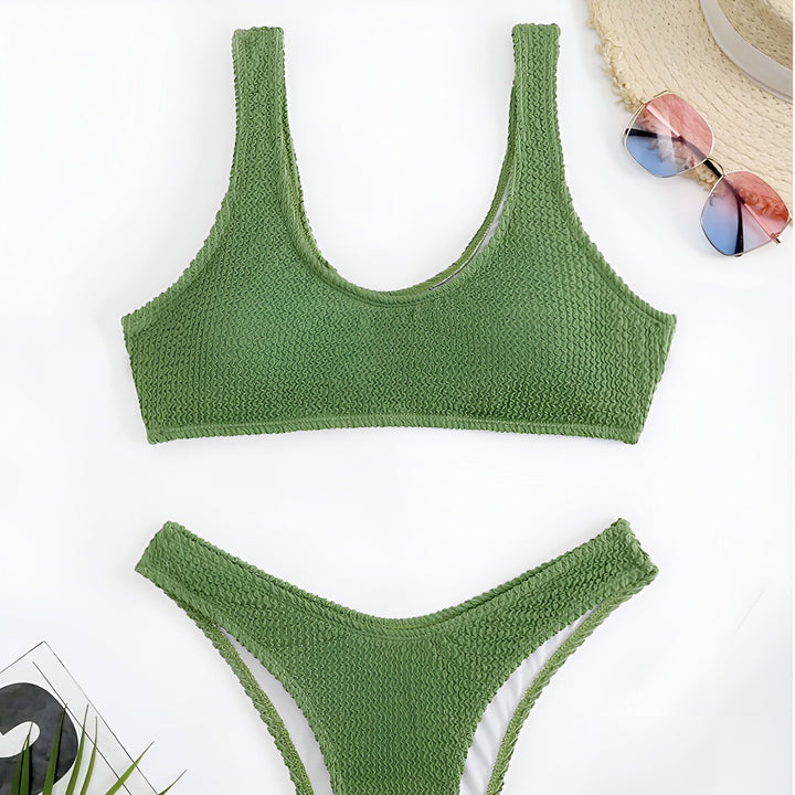 Femme en maillot de bain vert texturé, bronzée, assise sur une chaise longue. Détail du haut du bikini et de la peau. Mode estivale, vacances au soleil.