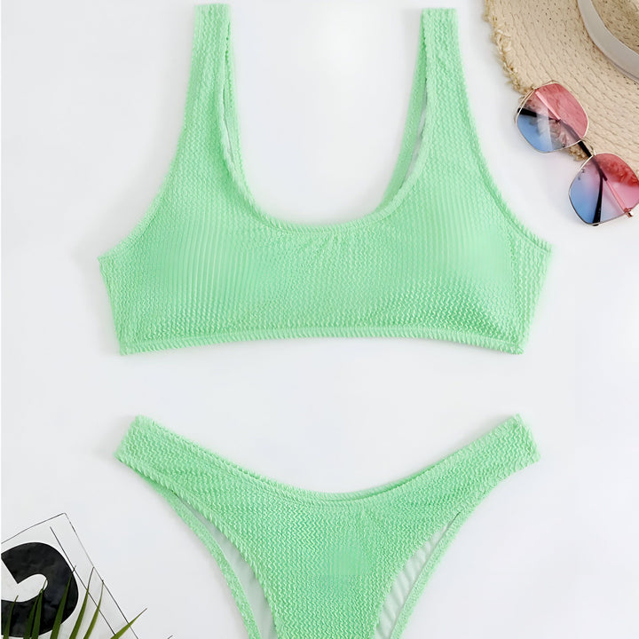 Vue aérienne d'un bikini vert citron texturé, un chapeau de paille, des lunettes de soleil multicolores et une plante, sur un fond blanc. Mode estivale.