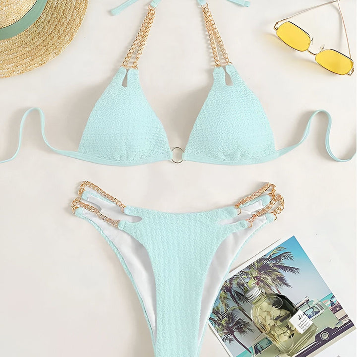 Femme en bikini rose texturé avec détails dorés, tenant un verre de vin rosé. Manucure bleue. Ambiance estivale et luxueuse.