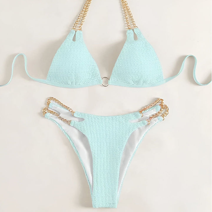 Image d'une femme portant un bikini texturé bleu clair avec des chaînes dorées, tenant un verre de vin rosé. Ses ongles sont vernis en bleu pastel.