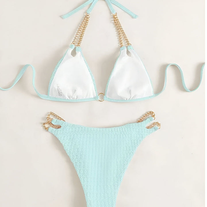 Femme en bikini rose texturé, tenant un verre de vin rosé. Détails de chaînes dorées sur le bikini. Manucure bleue visible. Mode estivale, vacances, détente.