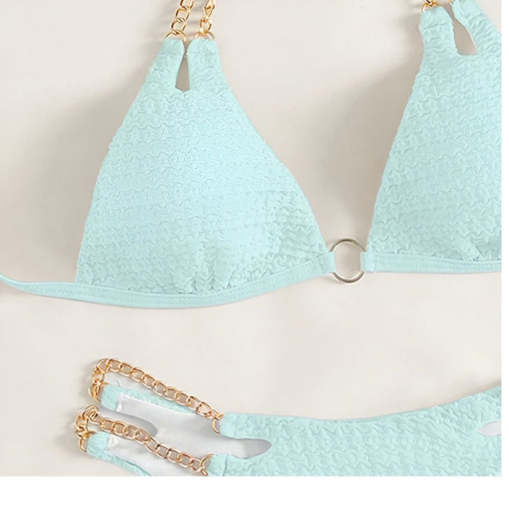 Femme en bikini jaune texturé avec détails en chaîne dorée tenant un verre de vin rosé. Manucure bleue assortie, bracelet doré. Élégance estivale, détente.