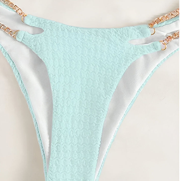 Femme en bikini vert pomme avec détails en chaîne dorée, tenant un verre de vin rosé. Manucure bleu clair. Mode estivale, vacances.