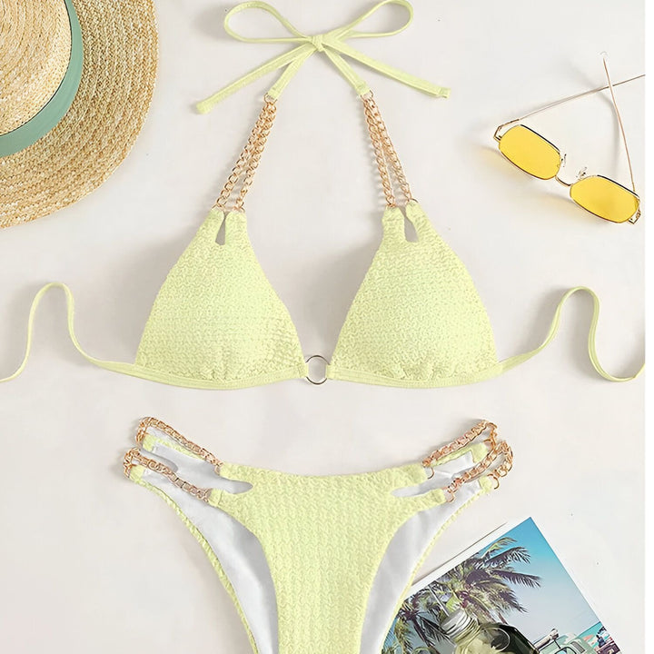 Image d'une femme en bikini noir texturé avec détails de chaîne dorée, tenant un verre d'eau. Manucure bleue pastel. Concept mode, été, plage, luxe.