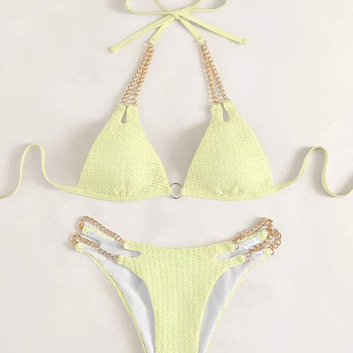 Femme en bikini bleu texturé avec des chaînes dorées, tenant un verre d'eau. Vernis à ongles bleu assorti.