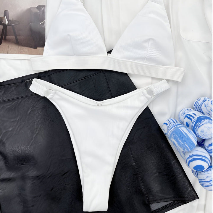 Image d'un bikini bleu électrique en côte rayée, composé d'un haut triangle et d'une culotte tanga avec anneaux, posé sur une jupe en similicuir noire et une chemise blanche, pour un style estival tendance.