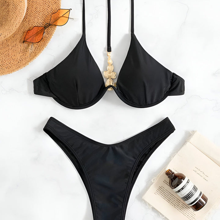 Image d'un ensemble de bikini noir, comprenant un haut orné et une culotte assortie. À côté, on voit un chapeau de paille, des lunettes de soleil originales, une bouteille cosmétique et un livre ouvert. Idéal pour l'été!