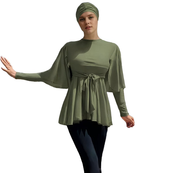 Femme portant un burkini vert olive avec un nœud à la taille et un legging noir sur une plage ensoleillée. Vêtements de bain islamiques modernes et élégants.