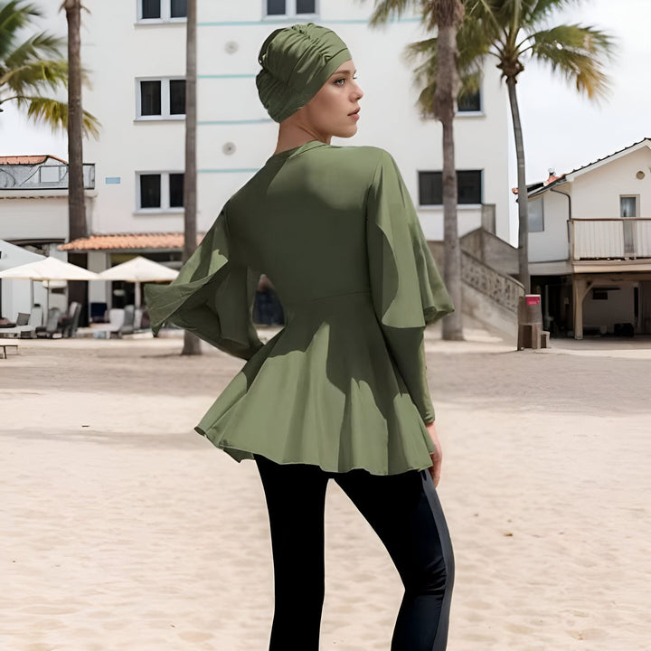 Femme portant un maillot de bain modeste olive avec un hijab assorti sur une plage ensoleillée avec des palmiers et un bâtiment blanc en arrière-plan.