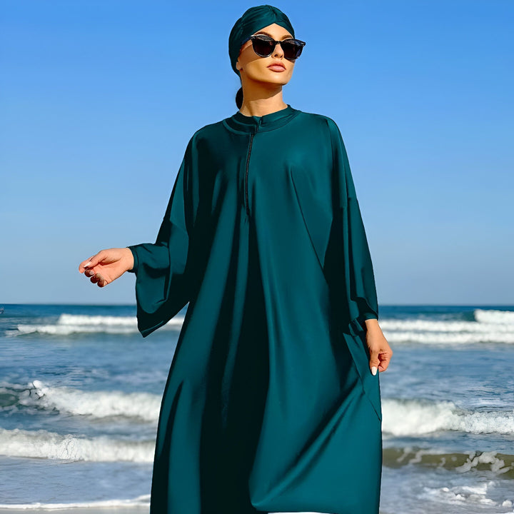 Femme portant un maillot de bain burkini vert foncé sur une plage ensoleillée. Le ciel bleu et les vagues de l'océan sont visibles en arrière-plan. Vêtements de bain islamiques.