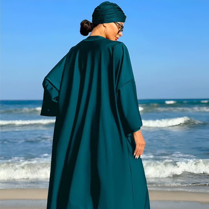 Femme portant un burkini vert émeraude avec un turban assorti et des lunettes de soleil, dos à la caméra, sur une plage ensoleillée avec l'océan en arrière-plan.