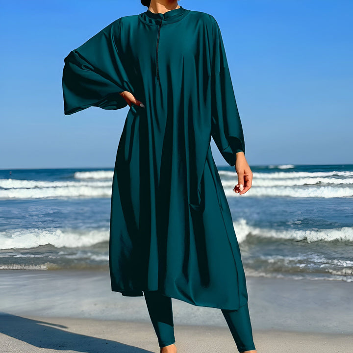 Femme en burkini turquoise et turban assorti, avec des lunettes de soleil, devant la mer et un ciel bleu clair. Mode islamique pudique sur la plage.