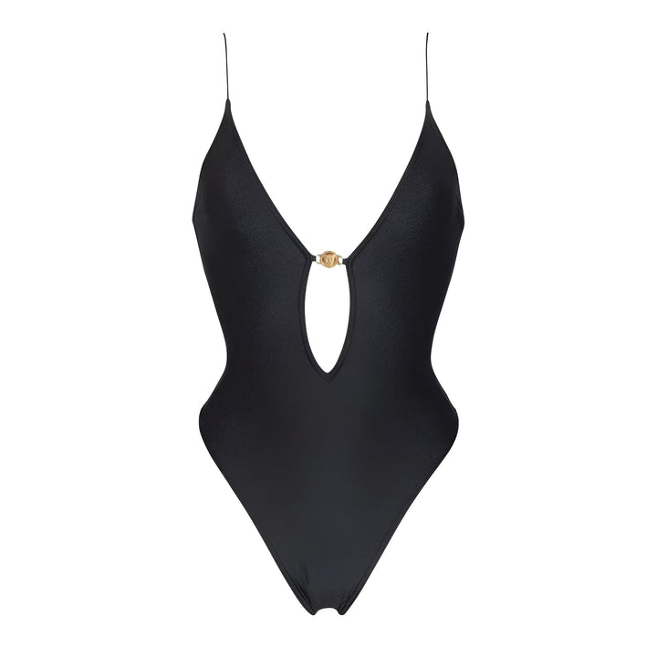 Une femme en maillot de bain noir une pièce à découpe, allongée près d'une table basse en bois rustique avec des bougies et un livre. Cheveux longs et châtains. Ambiance intérieure chaleureuse et décontractée.