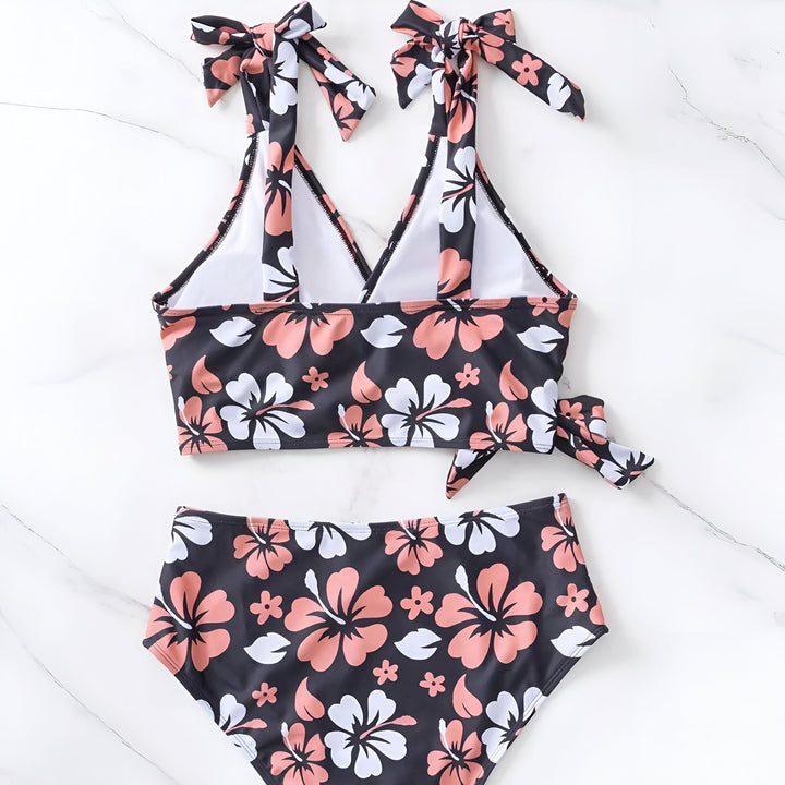 Maillot de bain deux pièces à motifs floraux sur fond noir, avec un chapeau de paille et des lunettes de soleil. Idéal pour la plage et les vacances d'été.