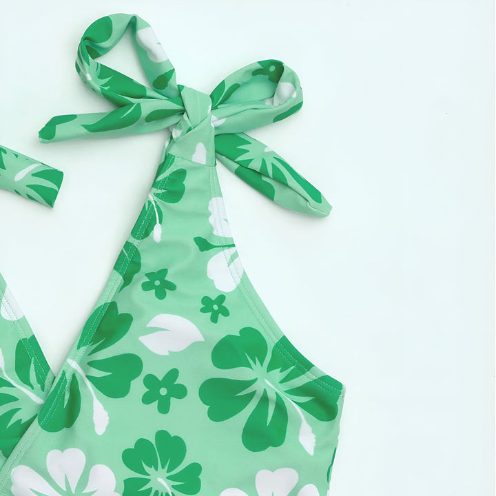 Dos de femme en maillot de bain violet à motifs floraux, portant un chapeau de paille. Vêtements posés à côté. Style estival, vacances.