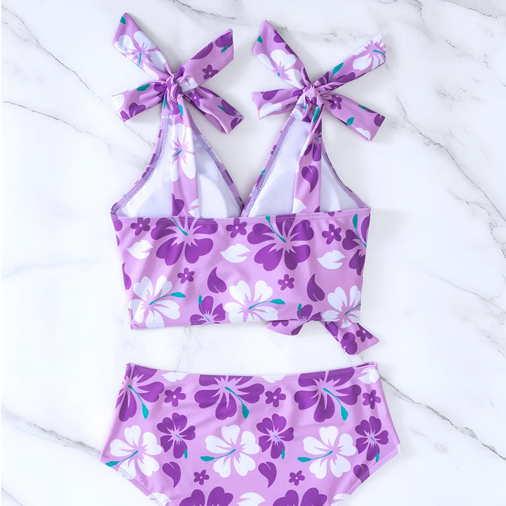 Femme portant un bikini orange à fleurs blanches et rouges. Le haut est noué sur l'épaule et sur le côté. Elle pose sa main sur sa hanche. Vêtement de bain tendance pour l'été.