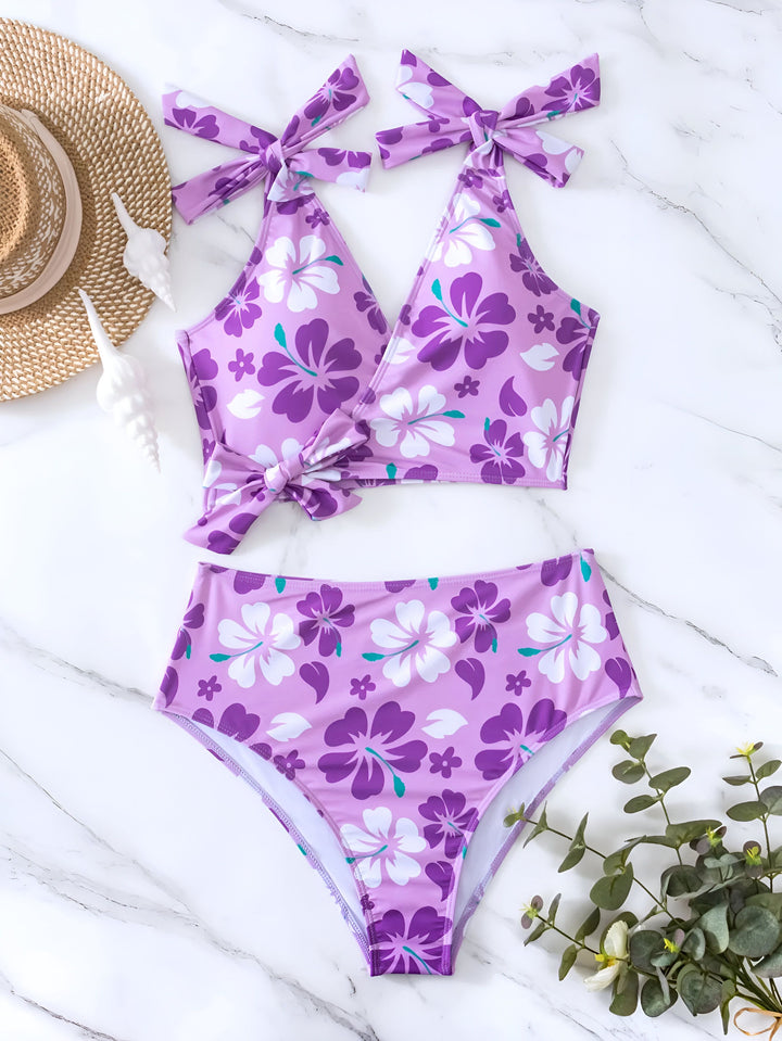 Femme portant un maillot de bain deux pièces orange à motifs floraux rouges et blancs, posant sur un fond neutre. Bretelles à nouer, taille haute.