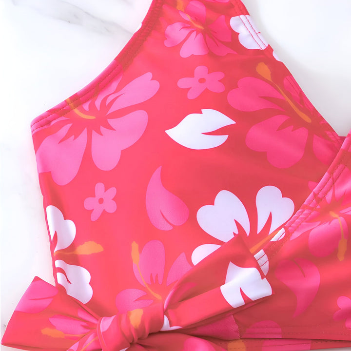 Image d'une femme en maillot de bain orange à motifs floraux, coiffée d'un chapeau de paille et portant une veste en jean claire. Elle est allongée sur un lit. Photographie de mode estivale, look décontracté et ensoleillé.