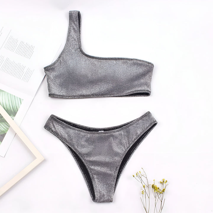 Une femme souriante en bikini scintillant gris est allongée sur un transat à la plage. On aperçoit le sable, la mer et une personne au loin.