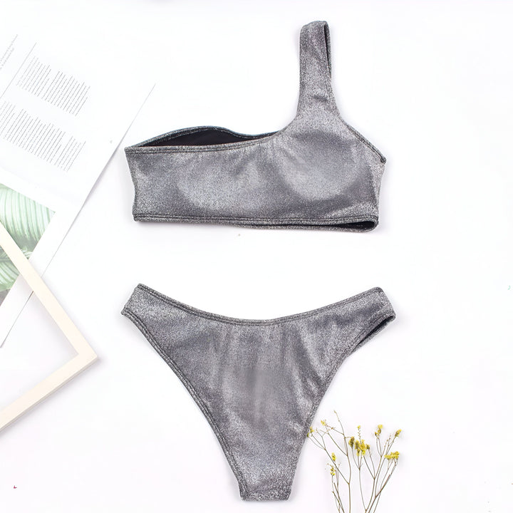 Image d'un maillot de bain deux pièces argenté pailleté avec un haut asymétrique et une culotte assortie, posé sur un fond blanc avec des accessoires décoratifs.