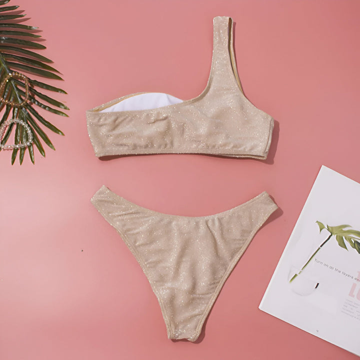 Ensemble de bikini doré scintillant une épaule sur fond rose, avec une feuille de palmier et des bracelets. Idéal pour l'été et la plage.