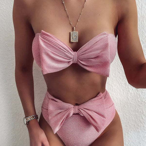 Femme portant un bikini rose pailleté avec des nœuds, et un collier doré avec un pendentif rectangulaire à motif floral.