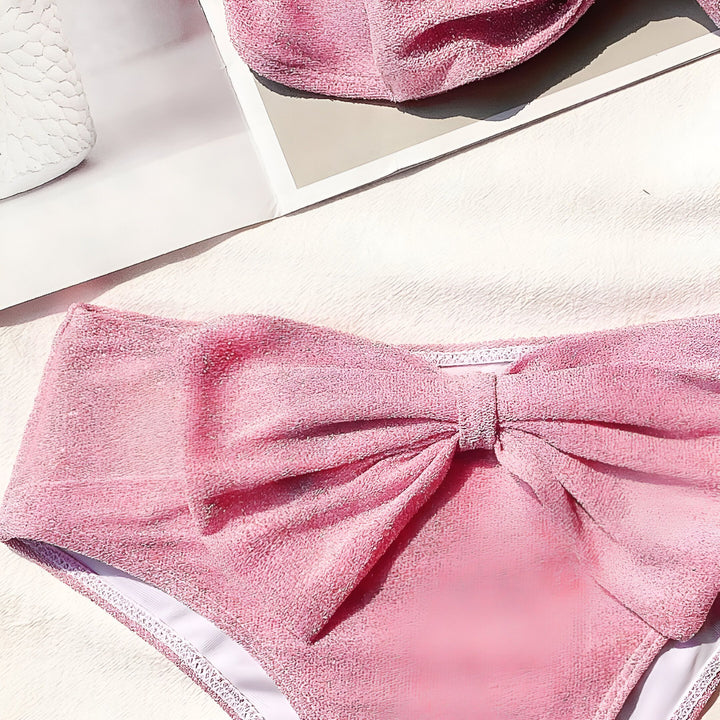 Culotte de bain rose texturée avec un grand nœud, faisant partie d'un ensemble avec un haut de bikini et un chapeau assortis. Mode estivale, vêtements de plage, style élégant et ludique.