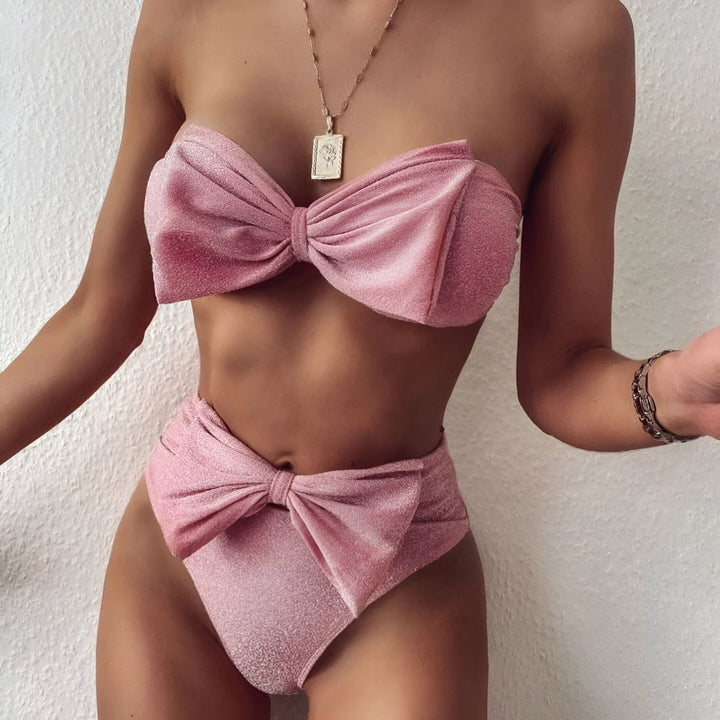 Femme en maillot de bain rose à paillettes avec des nœuds papillon, portant un collier avec un pendentif carré doré et une montre. Mode été, style élégant.