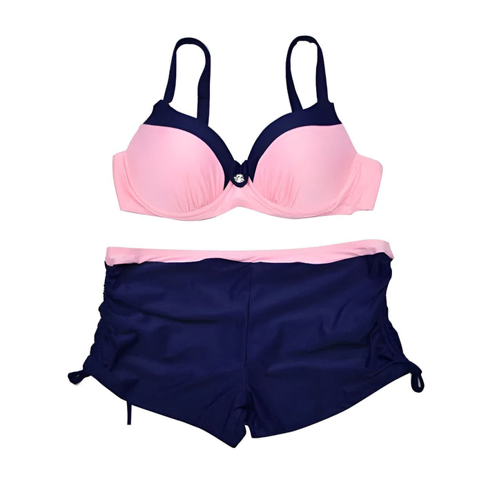Femme en maillot de bain deux pièces rose et bleu marine, posant devant un mur blanc. Haut de bikini rembourré avec détail bouton. Short de bain avec liens sur les côtés. Cheveux longs et tatouage sur le bras. Mode été, vêtements de plage.