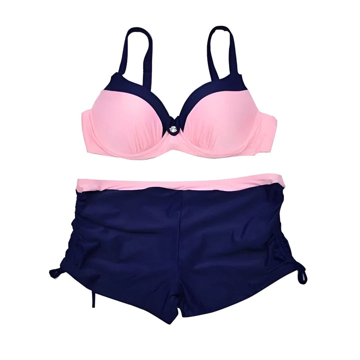 Femme en maillot de bain deux pièces rose et bleu marine. Haut de bikini à armatures avec détail bouton. Bas short de bain avec cordons sur les côtés. Bronzage, plage.