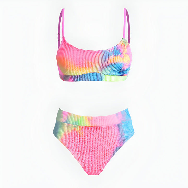 Femme blonde souriante en bikini multicolore sur une plage de sable fin. Elle porte un collier et des bracelets de coquillages. L'océan bleu turquoise est visible en arrière-plan. Ambiance estivale et ensoleillée.