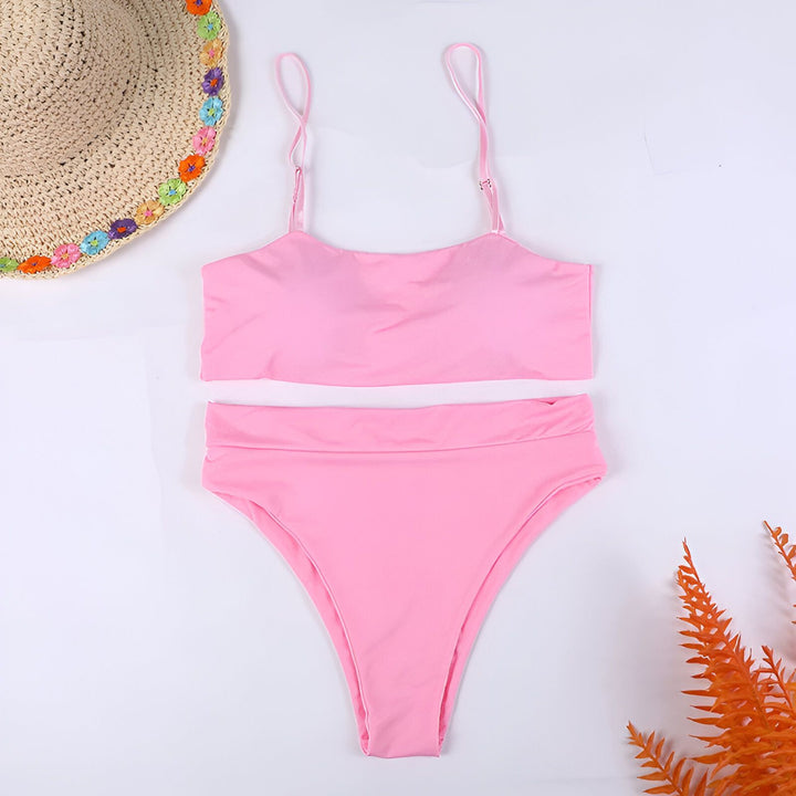 Femme portant un maillot de bain deux pièces rouge vif, avec un haut bandeau et une culotte taille haute, posant devant un mur blanc texturé. Lumière naturelle accentuant les contours du corps. Mode estivale, bronzage.