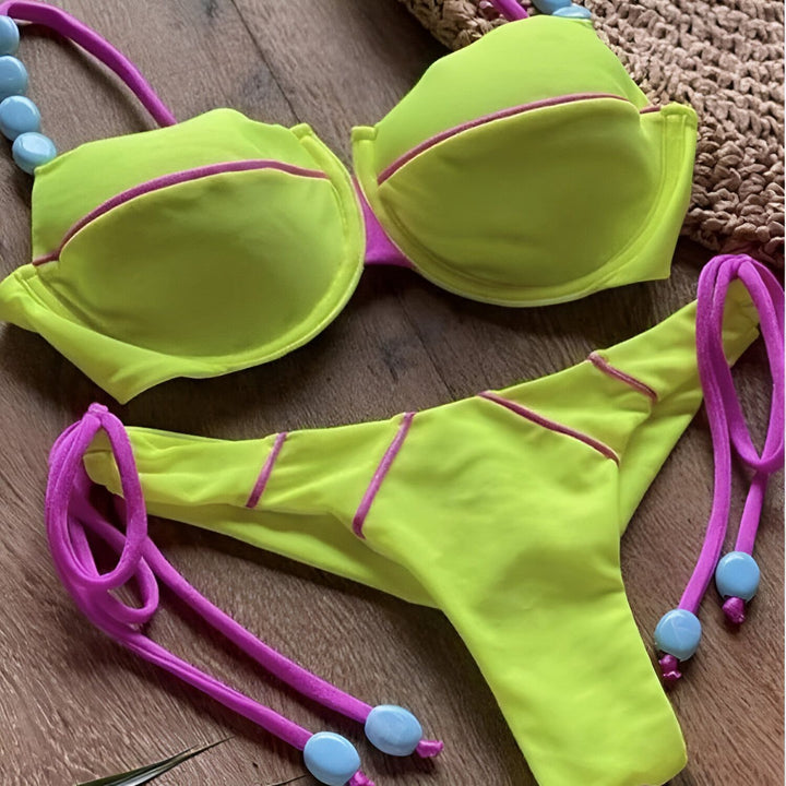 Image d'un bikini turquoise avec bordure violette, agrémenté de perles bleues, reposant sur une surface duveteuse. Un chapeau de paille à pois est également visible dans le cadre.