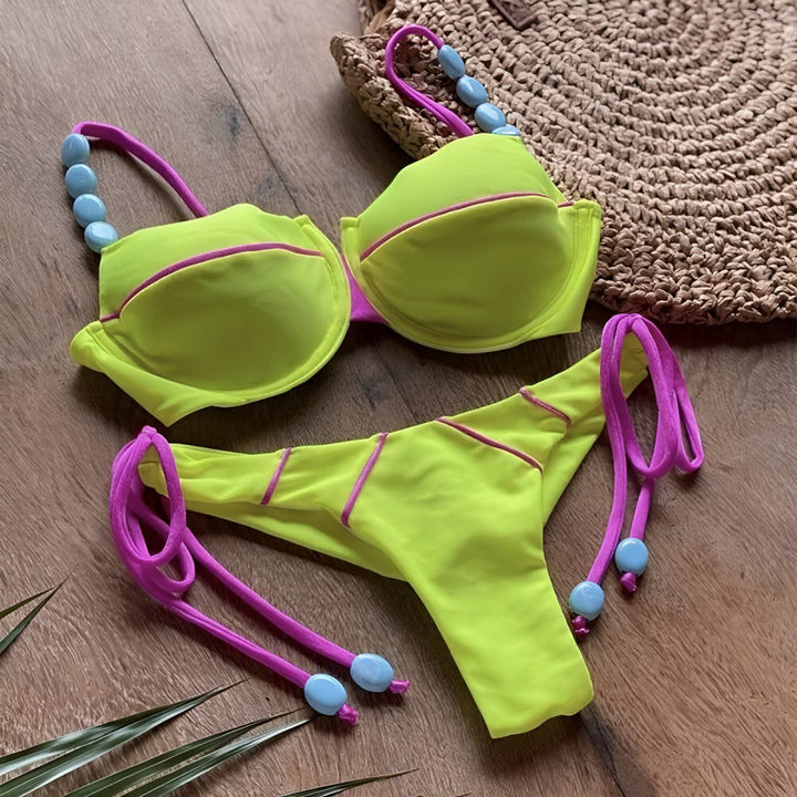Ensemble de bikini rose vif avec détails jaunes et cordons vert citron à perles bleues, posé sur une fourrure blanche avec un chapeau de paille en arrière-plan. Idéal pour la plage, l'été et les vacances.