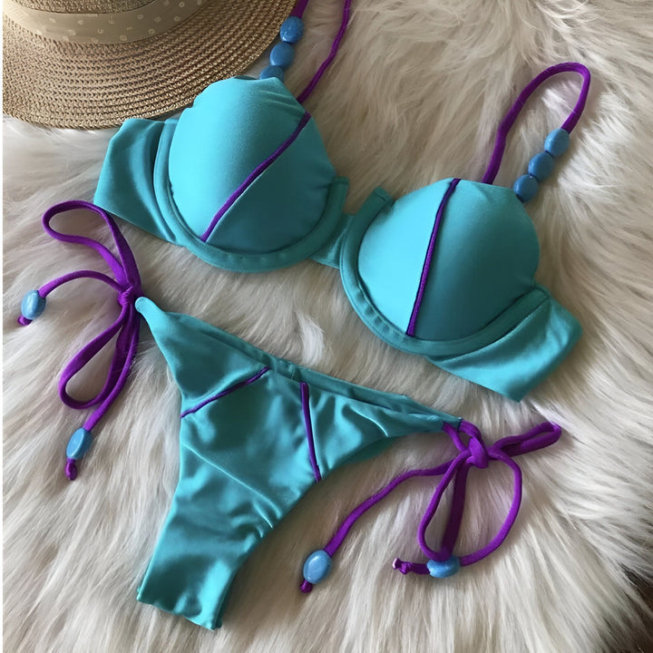 Image d'un bikini turquoise et violet avec des détails perlés, posé sur une surface duveteuse blanche. Un chapeau de paille est visible dans le coin supérieur gauche. Le bikini est composé d'un soutien-gorge et d'une culotte assortie.