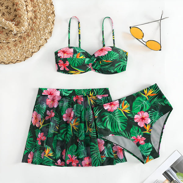 Une femme portant un bikini imprimé tropical noir, vert et rose, avec des feuilles de palmier et des fleurs d'hibiscus. Elle porte un bracelet doré. Le ciel bleu est en arrière-plan.