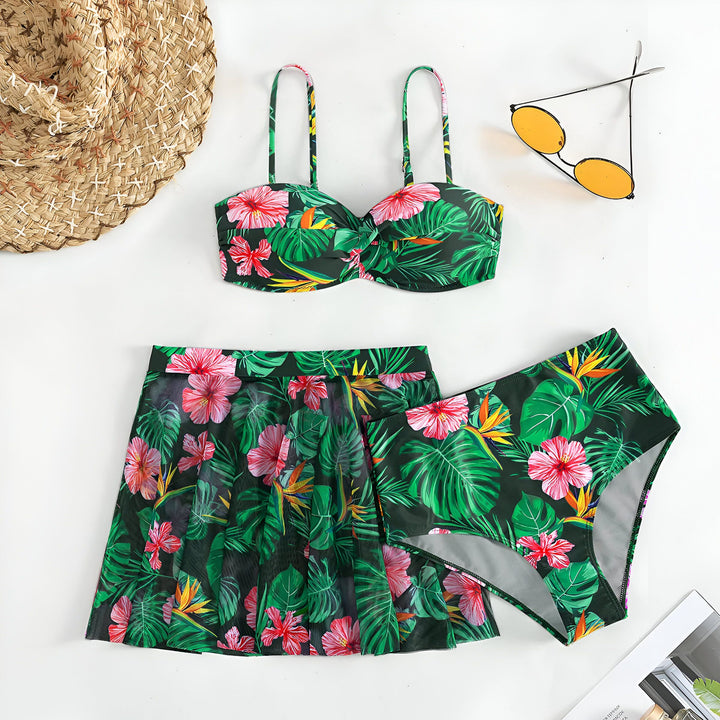 Une femme portant un bikini imprimé tropical noir, vert et rose, avec des feuilles de palmier et des fleurs d'hibiscus. Elle porte un bracelet doré. Le ciel bleu est en arrière-plan.