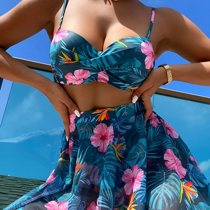 Femme portant un bikini et une jupe assortis à motifs floraux sur fond de ciel bleu. Tenue de plage tropicale, mode estivale.