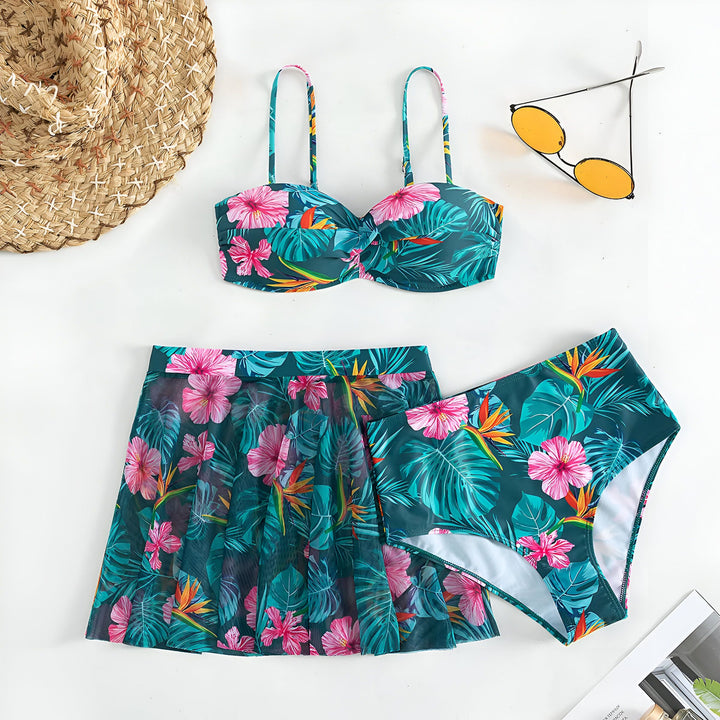 Ensemble de maillot de bain deux pièces bleu à imprimé floral tropical, comprenant un haut bandeau, une culotte taille haute et une jupe assortie. Chapeau de paille et lunettes de soleil jaunes complètent le look estival.