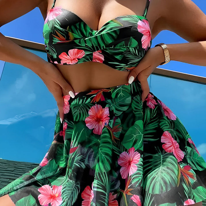 Ensemble de maillot de bain deux pièces avec imprimé floral tropical, comprenant un haut de bikini bandeau, une culotte de bain et une jupe assortie. Chapeau de paille et lunettes de soleil jaunes sur fond blanc.