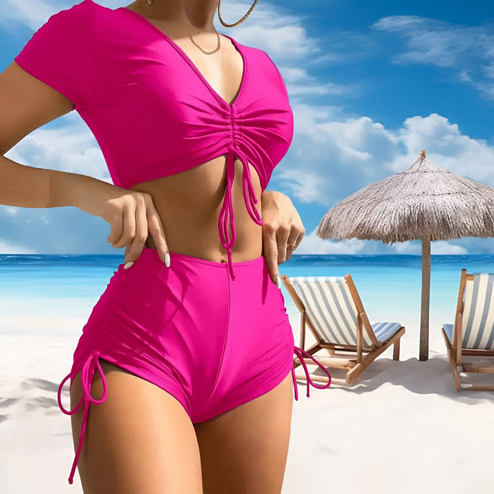 Une femme sur une plage porte un maillot de bain rose vif deux pièces avec des cordons de serrage, posant devant l'océan bleu, un parasol et des chaises longues.