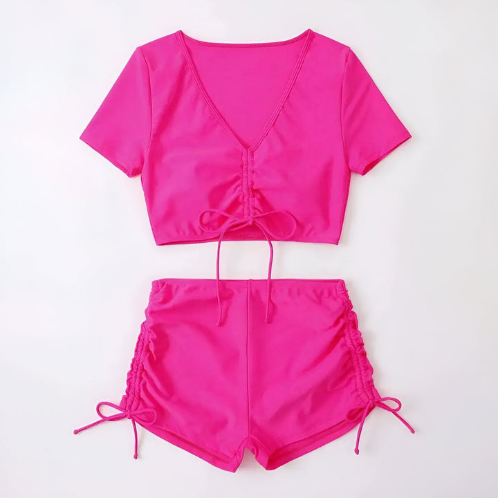 Femme portant un ensemble rose vif composé d'un haut noué court et d'un short de bain assorti, posant devant un mur texturé.