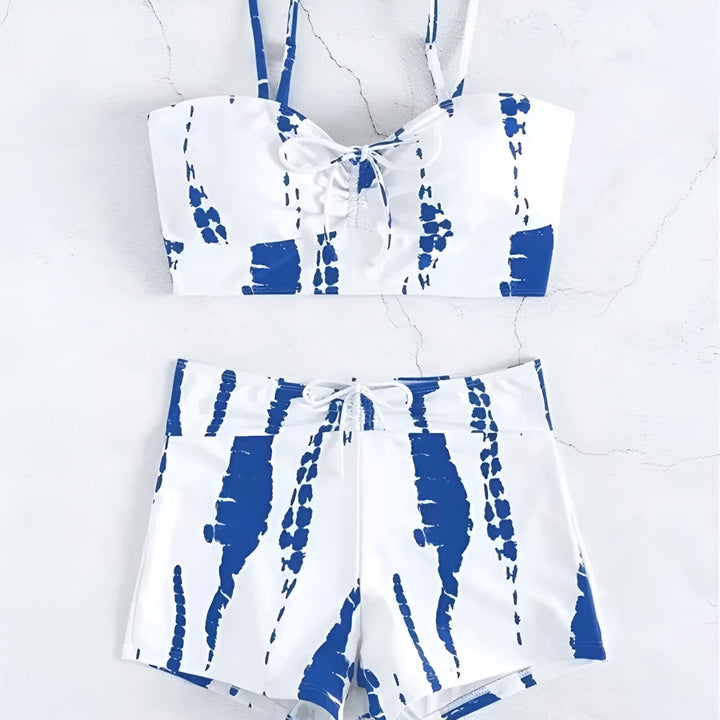 Femme blonde de dos en ensemble short et crop top blanc et marron au bord de la piscine. Détails de la décoration murale bleue et turquoise, ambiance estivale.